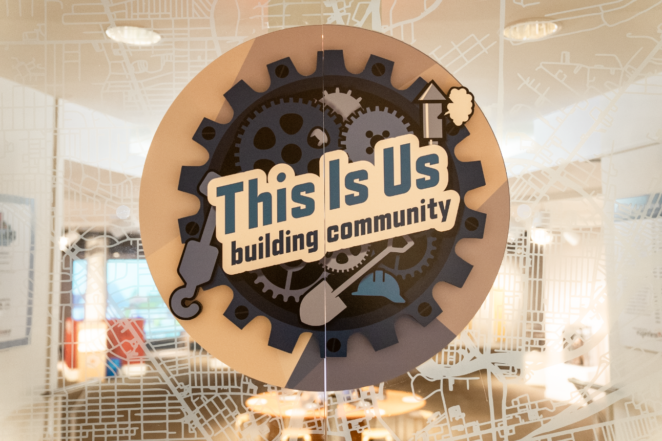 A close-up of a logo on the doors to the Lange Family Experience Center reading "This is Us" with :you, Me, & Dayton" written in smaller letters below. Above the words is a graphic rendition of the Dayton skyline featuring a smiling sun peaking around the tallest building.